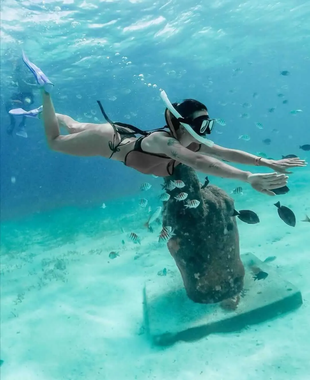 Tattooed snorkeler girl swimming among fish in front of underwater statue