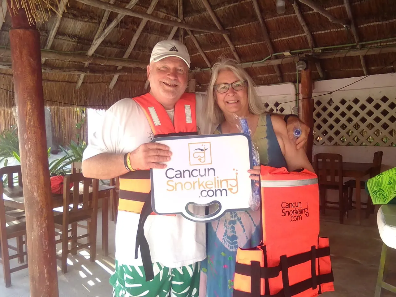 Adult couple at palapa roofed restaurant wearing life jackets and holding a sign with Cancun Snorkeling logo