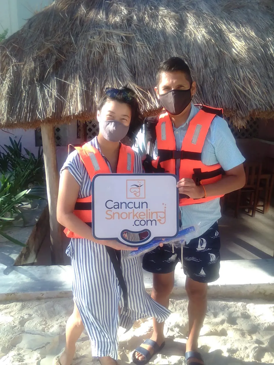 Young couple standing in the sand wearing life jackets and holding a sign that reads Cancun Snorkeling