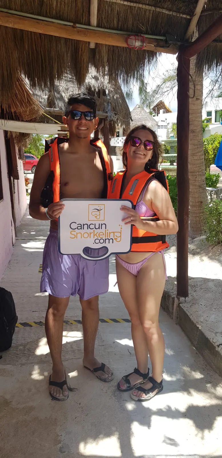 Young couple on swimsuits wearing life jackets and sunglasses holding a sign with the Cancun Snorkeling logo